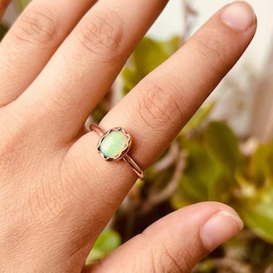 May include: A rose gold ring featuring an oval, light green opal gemstone with a scalloped bezel setting. The ring is displayed on a finger, showcasing the delicate design and color of the gemstone. The ring is a piece of jewelry.