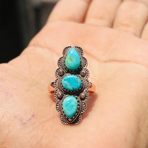 May include: A copper ring featuring three turquoise stones set in a vertical arrangement. The center stone is oval, flanked by two pear-shaped stones. The metal setting has a scalloped, stamped edge with a dark, oxidized finish around the stones.