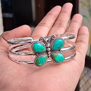 May include: A silver cuff bracelet featuring a central butterfly motif. The butterfly is composed of four teardrop-shaped turquoise stones with brown matrix veining, set in twisted silver wire borders. The band consists of three parallel silver wires.