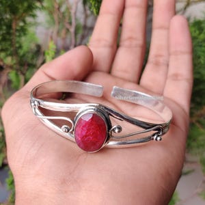 May include: A silver cuff bracelet featuring a large, oval-cut, deep red gemstone. The bracelet has an open design with decorative swirls and small ball accents. The text "SilverGemstoneRing" is visible on the bracelet.