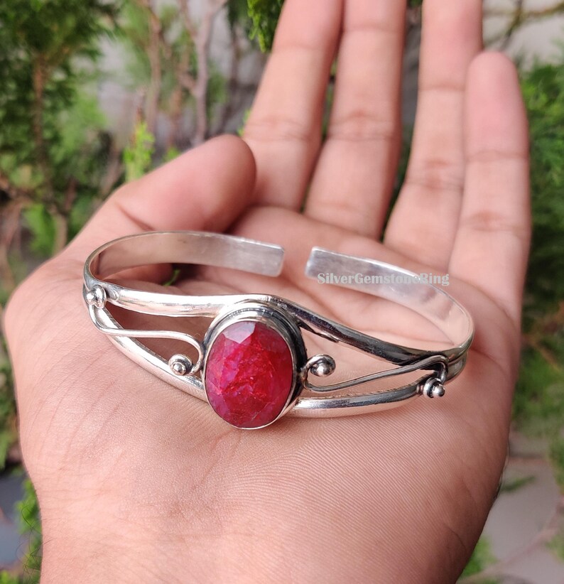 May include: A silver cuff bracelet featuring a large, oval-cut, deep red gemstone. The bracelet has an open design with decorative swirls and small ball accents. The text "SilverGemstoneRing" is visible on the bracelet.