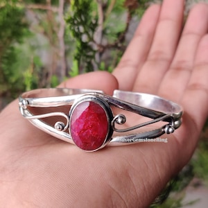 Könnte beinhalten: Ein silbernes Armreif-Armband mit einem ovalen, tiefroten Edelstein in der Mitte. Das Armband hat dekorative Wirbel und kleine Kugelakzente. Der Text "SilverGemstoneRing" ist sichtbar.