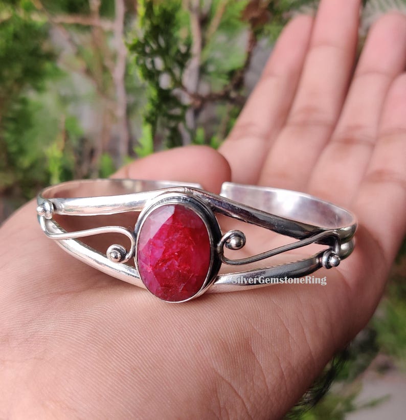 May include: A silver cuff bracelet featuring a large, oval, deep red gemstone. The bracelet has decorative swirls and small ball accents. The text "SilverGemstoneRing" is visible on the bracelet.
