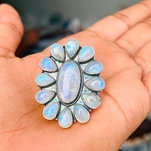 Puede incluir: Un anillo de plata con una gran piedra lunar ovalada central rodeada por doce piedras lunares m&aacute;s peque&ntilde;as en forma de l&aacute;grima dispuestas en un patr&oacute;n floral. Las piedras tienen un aspecto blanco lechoso transl&uacute;cido con destellos azules.