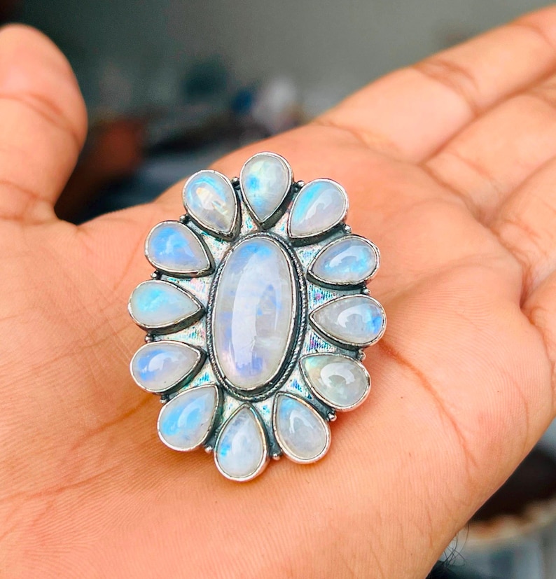 Puede incluir: Un anillo de plata con una gran piedra lunar ovalada central rodeada por doce piedras lunares m&aacute;s peque&ntilde;as en forma de l&aacute;grima dispuestas en un patr&oacute;n floral. Las piedras tienen un aspecto blanco lechoso transl&uacute;cido con destellos azules.