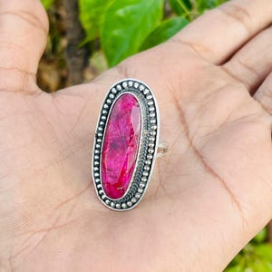 May include: A silver ring with an oval, vibrant pink gemstone. The gemstone is surrounded by a beaded border and set in a silver band. The ring is displayed on a hand.
