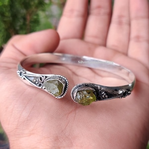 May include: A silver cuff bracelet with two olive-green gemstones. The open design bracelet has decorative detailing around the stones. The text "SilverGemstoneRings" is engraved on the bracelet.