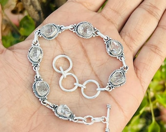 May include: A silver bracelet featuring clear, oval-shaped gemstones set in ornate bezels. The bracelet has a toggle clasp and is displayed on a hand against a backdrop of green foliage. The gemstones are approximately 0.5 inches in length.