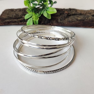 Pode incluir: Um conjunto de pulseiras de prata com diferentes designs. Algumas são lisas, enquanto outras apresentam padrões intrincados, como texturas florais ou trançadas. As pulseiras são exibidas em uma superfície branca, com um pedaço de madeira e folhas verdes ao fundo.
