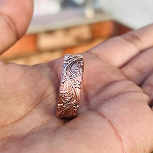 Peut inclure: Une bague de couleur cuivrée ornée d'un motif floral et de vigne complexe. La surface de la bague est recouverte de gravures détaillées de fleurs, de feuilles et de motifs tourbillonnants. La bague est de style alliance, avec une largeur constante.