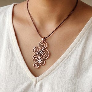 May include: A copper-colored pendant necklace featuring a cluster of spiral designs. The pendant hangs from a brown cord. The necklace is worn against a white shirt.