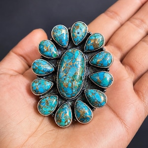 Puede incluir: Un anillo grande con una piedra turquesa ovalada central rodeada por once piedras turquesas m&aacute;s peque&ntilde;as en forma de pera. Cada piedra muestra un color azul vibrante con vetas de matriz de color cobre, engastadas en un marco de metal plateado.