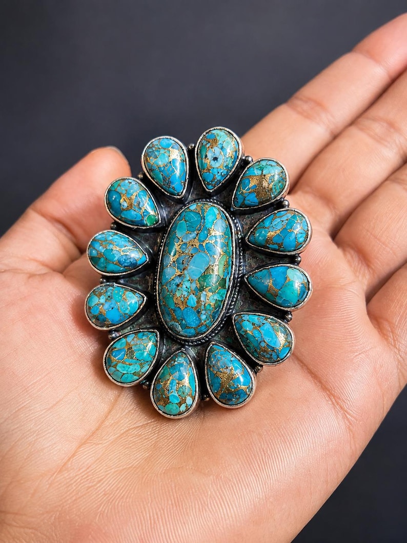 Puede incluir: Un anillo grande con una piedra turquesa ovalada central rodeada por once piedras turquesas m&aacute;s peque&ntilde;as en forma de pera. Cada piedra muestra un color azul vibrante con vetas de matriz de color cobre, engastadas en un marco de metal plateado.