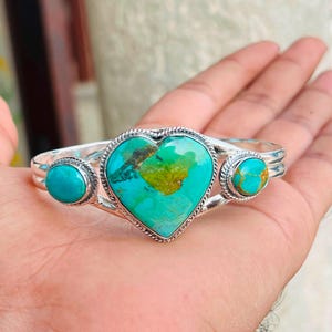 May include: A silver cuff bracelet featuring a central heart-shaped turquoise stone flanked by two smaller round turquoise stones. The metal setting has a twisted rope texture around each stone, and the band is composed of two parallel silver wires.