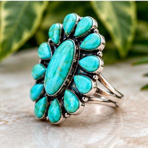 Puede incluir: Un anillo de plata con una gran piedra turquesa ovalada central rodeada por doce piedras turquesas m&aacute;s peque&ntilde;as en forma de l&aacute;grima. Las piedras est&aacute;n engastadas en biseles de plata pulida con un dise&ntilde;o de banda de v&aacute;stago dividido.