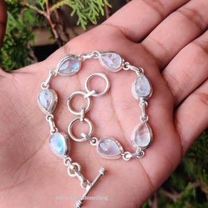 May include: A silver bracelet with pear-shaped moonstone gemstones. The bracelet has a toggle clasp and is displayed on a hand. The moonstones have a blue and white iridescent glow. The bracelet is connected by small silver links.