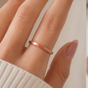 May include: A simple, smooth, polished copper-toned metal band ring with a flat profile and a consistent width, worn on a finger. The ring features a warm, metallic finish that reflects light evenly across its surface, showcasing a clean design.