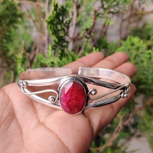 May include: A silver cuff bracelet with a large, oval, bright red gemstone. The bracelet has an open design with decorative swirls and small silver accents. The background is a blurred view of green foliage.
