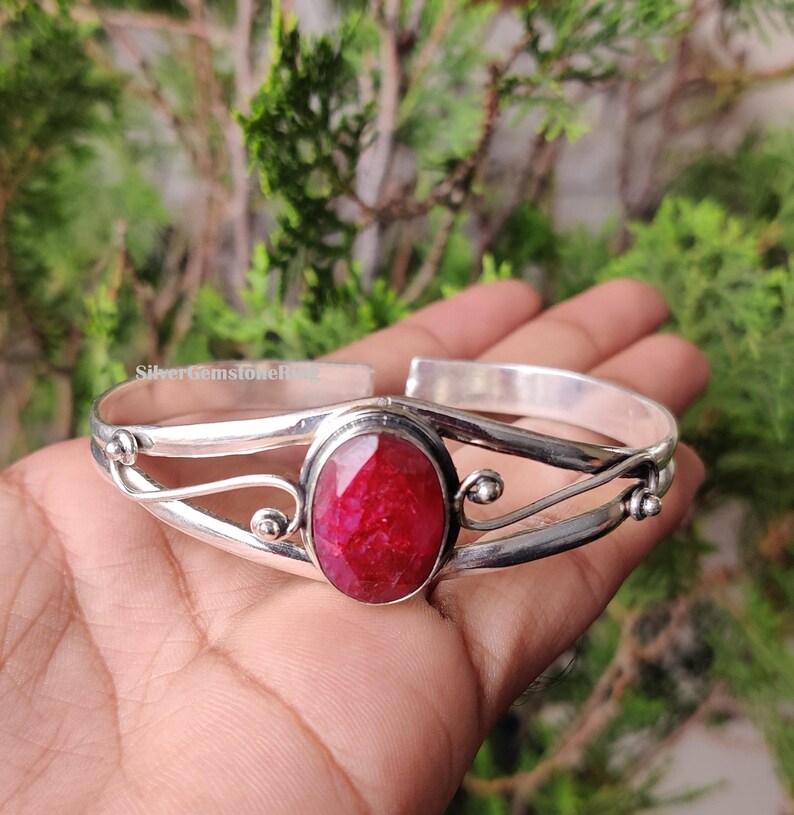 May include: A silver cuff bracelet with a large, oval, bright red gemstone. The bracelet has an open design with decorative swirls and small silver accents. The background is a blurred view of green foliage.