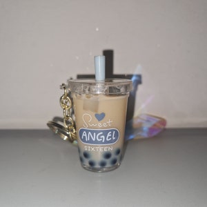 Può includere: Un portachiavi in plastica trasparente a forma di bicchiere di boba tea con una cannuccia blu e bianca. Il bicchiere contiene un liquido marrone e perle di boba blu. Sul bicchiere è stampato il testo "Sweet Angel Sixteen".