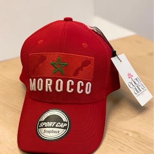 Peut inclure: Casquette de sport rouge avec le mot "MOROCCO" brodé en blanc. La casquette présente un écusson avec une étoile verte et le contour du Maroc. Une étiquette avec les mots "eden aura" est accrochée à la casquette.