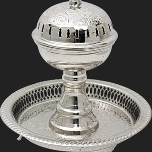 May include: A silver-colored incense burner with a domed lid and a decorative tray base. The lid has small cut-out vents and a small knob on top. The tray has a decorative pattern around the edge and is supported by three feet.