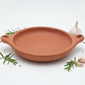 May include: A terracotta serving dish with two handles, surrounded by sprigs of rosemary, peppercorns, garlic cloves, and salt. The dish is a warm, earthy tone, perfect for presenting food.
