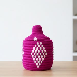 Peut inclure: Un pot de rangement tissé magenta avec un couvercle. Le pot a un corps cylindrique et un dessus arrondi. Un motif de losange blanc décore le devant. Le pot est fait d'un matériau épais et texturé et repose sur une surface en bois.