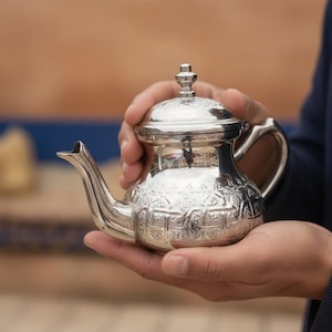 Peut inclure: Une théière argentée avec des motifs en relief complexes est tenue dans deux mains. La théière a un bec courbé, une anse et un couvercle avec un petit bouton. La théière est probablement en métal et reflète la lumière.