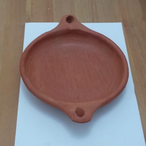 May include: A terracotta-colored, oval-shaped dish with two handles, each featuring a hole. The dish is placed on a white surface, set against a wooden background. The design is simple and rustic.