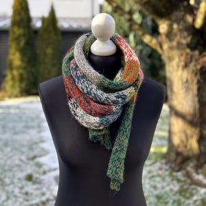 Könnte beinhalten: Ein mehrfarbiger Strickschal, präsentiert auf einer schwarzen Schaufensterpuppe. Der Schal weist eine Mischung aus grünen, orangefarbenen, grauen und blauen Farbtönen auf. Er besteht aus dickem Garn und hat eine strukturierte Optik. Der Schal hat eine dreieckige Form.