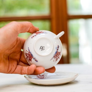 Op de afbeelding: Een wit theekopje met een rood en blauw bloemenmotief. De onderkant van het kopje heeft een zwarte markering met de tekst "F.T."