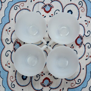 May include: Four white ceramic cups with a rounded shape and a smooth, glossy finish. The cups are arranged in a square pattern on a white tablecloth with a blue and red floral design.