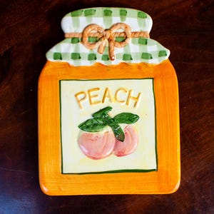 Könnte beinhalten: Ein dekorativer Teller aus Keramik zum Thema Pfirsich. Der Teller hat die Form eines Glases mit einem orangefarbenen Rahmen und einem grün-weiß karierten Deckel. Das Wort "PEACH" ist auf der Vorderseite zusammen mit zwei Pfirsich-Illustrationen geschrieben.