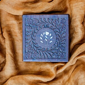 Può includere: Una scatola di legno con intagli intricati e un motivo floreale. La scatola è dipinta di un colore blu scuro e ha un'intarsio floreale bianco al centro.