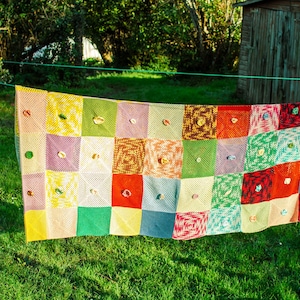 Könnte beinhalten: Eine bunte gehäkelte Decke mit Patchwork-Muster hängt im Freien an einer Wäscheleine. Die Decke besteht aus Quadraten in verschiedenen Farben, darunter Rot, Grün, Gelb, Blau und Lila.