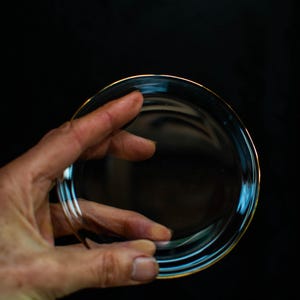 May include: A clear, round glass plate with a thin gold rim, held by a hand against a black background. The plate's transparent design allows light to pass through, creating reflections.