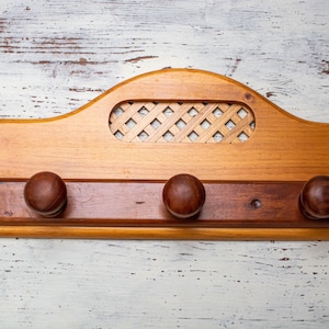 Może przedstawiać: Drewniany wieszak na ubrania z trzema haczykami i ozdobnym wzorem kratki. Wieszak wykonany jest z jasnego drewna brązowego i ma ciemnobrązowe wykończenie na haczykach.