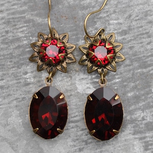 Puede incluir: Un par de pendientes colgantes con un engaste de metal en tono latón, con una parte superior en forma de flor que sostiene un cristal rojo redondo y facetado. Debajo, un cristal rojo oscuro ovalado cuelga del elemento floral mediante una anilla.