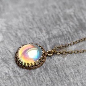 May include: A round pendant necklace with a shimmering, iridescent center. The pendant is set in a bronze-colored, ornate frame and hangs from a delicate chain. The necklace is displayed on a gray, textured surface.