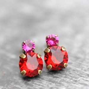 May include: A pair of stud earrings featuring a large, faceted red gemstone beneath a smaller, pink gemstone. The earrings have a gold-colored setting and are displayed against a gray background.