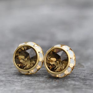 May include: A pair of gold-toned stud earrings. Each earring features a large, faceted, brown crystal center, surrounded by a ring of small, white, iridescent stones. The earrings are set against a gray background.