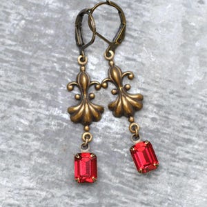 May include: A pair of antique gold-coloured earrings with a fleur-de-lis design. Each earring features a rectangular, vibrant red gemstone. The earrings have a lever-back closure and are designed for pierced ears.