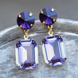 May include: A pair of earrings with large, rectangular, purple crystals hanging from smaller, round, dark purple crystals. The crystals are set in gold-coloured metal. The earrings are displayed on a textured, grey surface.