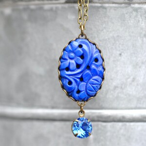 K&ouml;nnte beinhalten: Eine blaue, geschnitzte Anh&auml;nger-Halskette mit einem kleineren blauen Kristall-Anh&auml;nger. Der Anh&auml;nger zeigt ein florales Design und ist in einen Messingrahmen gefasst.