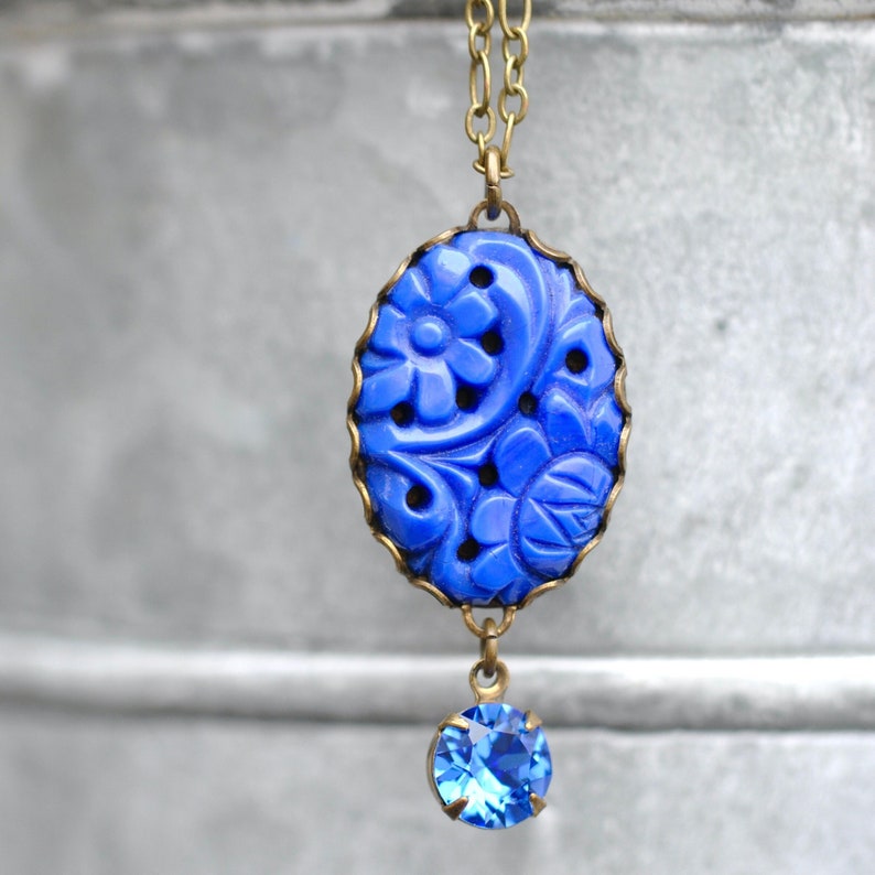 K&ouml;nnte beinhalten: Eine blaue, geschnitzte Anh&auml;nger-Halskette mit einem kleineren blauen Kristall-Anh&auml;nger. Der Anh&auml;nger zeigt ein florales Design und ist in einen Messingrahmen gefasst.