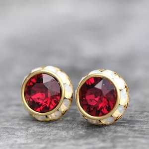 Op de afbeelding: Een paar oorstekers met grote, gefacetteerde, dieprode edelstenen gezet in goudkleurig metaal. De edelstenen zijn omgeven door een halo van kleine, witte, iriserende stenen.