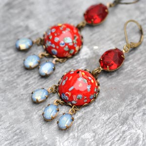 Op de afbeelding: Een paar rode en blauwe oorhangers met een ronde rode cabochon en blauwe kristallen accenten.