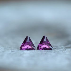 May include: Two triangular, faceted, deep purple gemstones. The gems are set against a blurred, light gray background. The facets catch the light, creating a sparkling effect. These gems could be used for jewelry making.