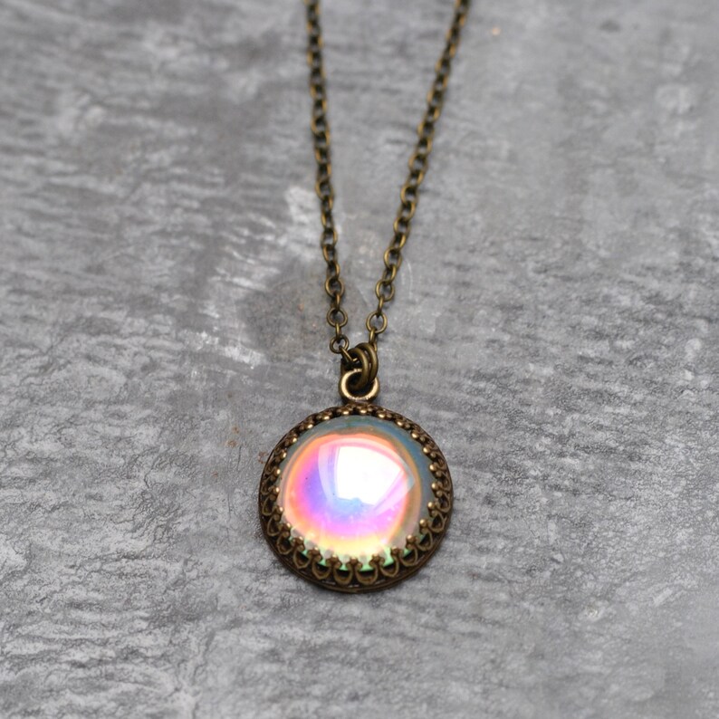 May include: A vintage-style necklace with a round pendant. The pendant features an iridescent, multi-colored stone set in a bronze-toned, filigree frame. The chain is also bronze-toned and hangs against a gray background.